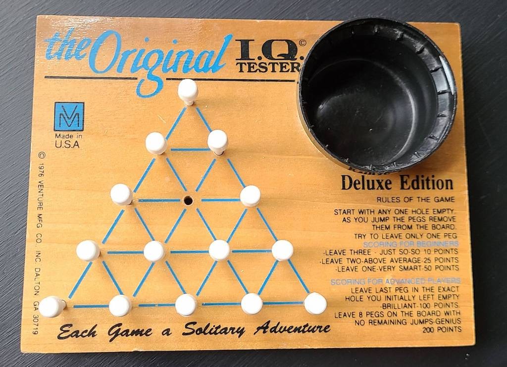 The Original I.Q. Tester Deluxe Edition game board — triangular peg layout with 14 pegs and one empty hole.